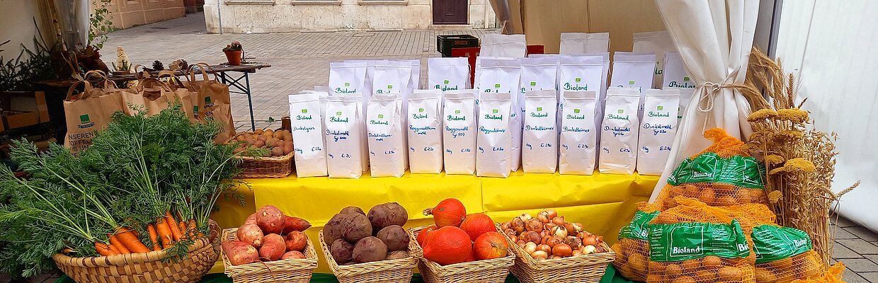Unter einem weißen Gartenzeltpavillon ist ein Biertisch mit einem gelben Tuch eingedeckt. Darauf stehen viele weiße Mehltüten. daneben ein Korb mit Kartoffeln, daneben braune Papiertüten mit Kartoffeln. Vor dem Tisch steht eine mit einem grünen Tuch und weißer Schrfit eingedeckte Bierbank. Darauf befinden sich nebeneinander füng verschiedene Körbe mit Bundkarotten, Süßkartoffeln, Rote Rüben, Hokkaido-Kürbisse und Zwiebeln, auch mehrere große Säcke Kartoffeln.
