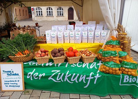 Unter einem weißen Gartenzeltpavillon ist ein Biertisch mit einem gelben Tuch eingedeckt. Darauf stehen viele weiße Mehltüten. daneben ein Korb mit Kartoffeln, daneben braune Papiertüten mit Kartoffeln. Vor dem Tisch steht eine mit einem grünen Tuch und weißer Schrfit eingedeckte Bierbank. Darauf befinden sich nebeneinander füng verschiedene Körbe mit Bundkarotten, Süßkartoffeln, Rote Rüben, Hokkaido-Kürbisse und Zwiebeln, auch mehrere große Säcke Kartoffeln.