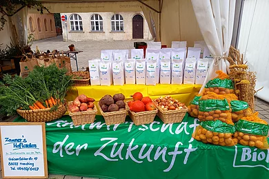 Unter einem weißen Gartenzeltpavillon ist ein Biertisch mit einem gelben Tuch eingedeckt. Darauf stehen viele weiße Mehltüten. daneben ein Korb mit Kartoffeln, daneben braune Papiertüten mit Kartoffeln. Vor dem Tisch steht eine mit einem grünen Tuch und weißer Schrfit eingedeckte Bierbank. Darauf befinden sich nebeneinander füng verschiedene Körbe mit Bundkarotten, Süßkartoffeln, Rote Rüben, Hokkaido-Kürbisse und Zwiebeln, auch mehrere große Säcke Kartoffeln.