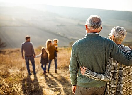 Ein älteres Paar, Mann und Frau, haben jeweils den Arm um den anderen gelegt, sind nur von hinten fotografiert und sehen, auf einem Trockenhang stehend ins Tal. Vor Ihnen befindet sich eine Familie, Mann, Frau und zwei Kinder.