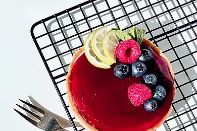 Ein kleiner Kuchen mit roter Glasur, garniert mit Beeren und Zitronenscheiben, auf einem Gitterrost.