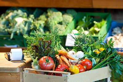 Holzkiste mit frischem Gemüse wie Tomaten, Karotten, Zwiebeln und Kräutern auf einem Marktstand.
