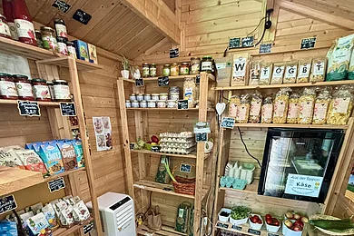 Kleiner Holzladen mit Regalen voller Lebensmittel, Eiern, Obst, Gemüse und Getränken.
