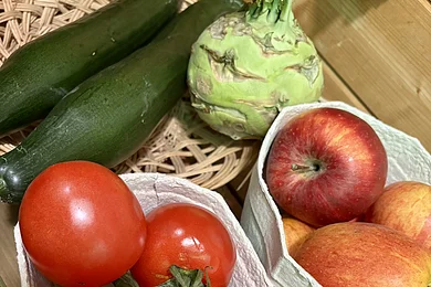 Tomaten, Äpfel, Gurken und Kohlrabi liegen auf einem Holztisch in Behältern und einem Korb.