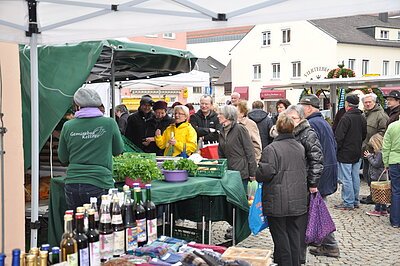 Gemüsestand mit Verkäuferin im grünen Pullover von hinten. Daneben stehen verschiedene Obstsaftflaschen. Der Stand steht auf dem Dorfplatz, umgeben von einer größeren Personengruppe. Im Hintergrund sieht man noch andere Marktstände.