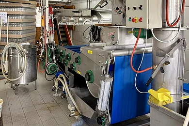 Industrielle Maschine mit Steuerung zum Entsaften von Obst. Rohre und Förderband in einem Raum mit Fliesenboden und -wänden.