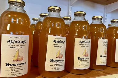 Mehrere Flaschen naturtrüber Apfelsaft mit goldfarbenen Deckeln auf einem Holzregal.